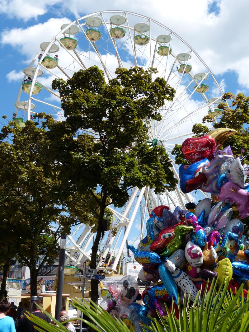 Riesenrad hinter grünen Bäumen bei blauem Himmel mit weißen Wolken, bunte Luftballons im Vordergrund