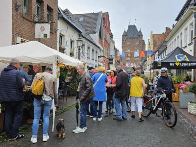 Menschen auf einer belebten Straße mit Marktständen und historischen Gebäuden im Hintergrund, darunter ein Tor mit Türmen