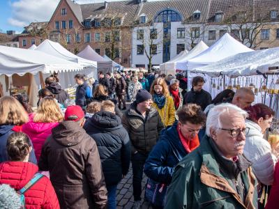 Menschenmenge auf einem belebten Markt mit weißen Verkaufszelten in einer Stadt bei klarem Himmel