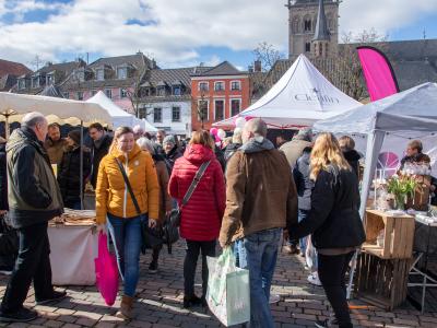 Menschen besuchen einen belebten Markt mit weißen Zelten und Ständen auf einem gepflasterten Platz vor historischen Gebäuden und einer Kirche