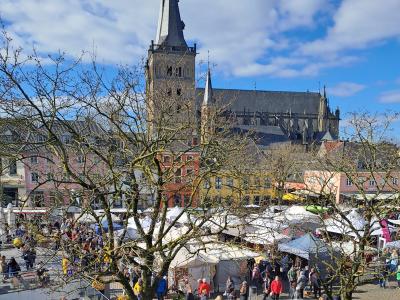 Weitwinkelaufnahme des belebten Marktplatzes von Xanrten in einer historischen Altstadt, überragt vom imposanten Dom mit gotischer Architektur. Zahlreiche Marktstände mit weißen Zeltdächern, viele Besucherinnen und Besucher sowie bunte Häuserfassaden im Hintergrund vermitteln eine lebendige Frühlingsatmosphäre unter blauem Himmel.