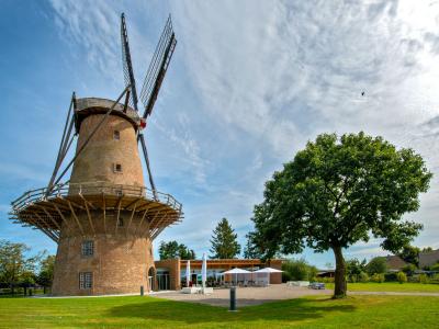 Ein traditioneller Windmühlenturm aus Backstein mit einem umlaufenden Balkon und schräg stehenden Flügeln vor einem blauen Himmel mit leichten Wolken. Rechts neben der Mühle steht ein großer, grüner Baum. Im Hintergrund befindet sich ein modernes Flachbaugebäude mit Außensitzplätzen. Die Szene wirkt ländlich und ruhig.