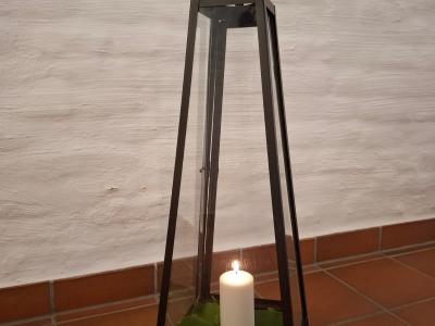 Ein Windlicht mit brennender Kerze. Das Windlicht steht vor einer Weißen Wand auf einem Terracottafarbenen Fliesenboden