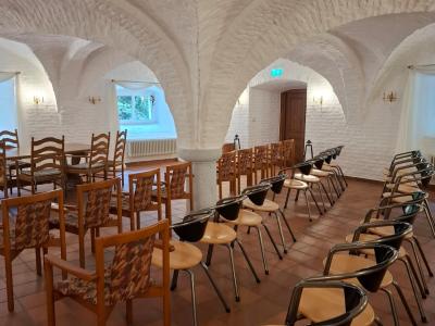 Ein stilvoll eingerichteter Raum mit weißen, gewölbten Wänden und Terrakotta-Fliesenboden. Links im Bild steht ein rechteckiger Holztisch mit fünf Stühlen im Landhausstil. Dahinter stehen drei Reihen mit Stühlen. Drei Fenster mit weißen Gardinen lassen natürliches Licht herein und geben den Blick auf grünes Laub frei. An den Wänden befinden sich dekorative Wandlampen. 