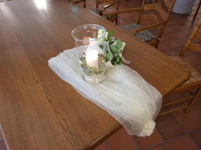 Auf einem Holztisch liegt eine zarte, weiße Stoffdekoration mit einer Kerze und Blumenschmuck. 