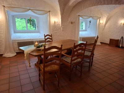 Ein stilvoll eingerichteter Raum mit weißen, gewölbten Wänden und Terrakotta-Fliesenboden. In der Mitte steht ein rechteckiger Holztisch mit sechs Stühlen im Landhausstil. Auf dem Tisch liegt eine zarte, weiße Stoffdekoration mit einer Kerze und Blumenschmuck. Zwei Fenster mit weißen Gardinen lassen natürliches Licht herein und geben den Blick auf grünes Laub frei. An den Wänden befinden sich dekorative Wandlampen. Die Atmosphäre ist ruhig und elegant.