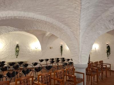 Ein stilvoll eingerichteter Raum mit weißen, gewölbten Wänden und Terrakotta-Fliesenboden. In der Mitte steht vier Reihen mit Stühlen, auf dem die Gäste platz finden. und Blumenschmuck. An den Wänden befinden sich dekorative Wandlampen.