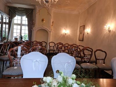 Auf dem Foto sieht man den Rokokosaal mit Ofennische. Vorne im Bild liegt auf dem Tisch ein Blumengesteck und dahinter stehen die weiß behussten Stühle für das Brautpaar. Im Hintergrund sind die Stühle für die Gäste aufgestellt. 