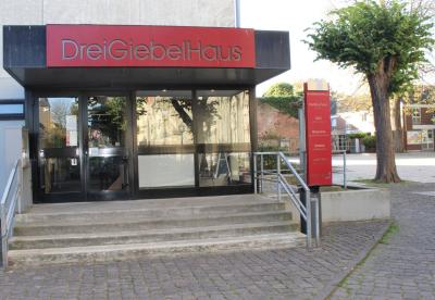 Der Haupteingang des DreiGiebelHaus von draußen. Über der schwarzen Glastür hängt ein Rotes Schild auf dem "DreiGiebelHaus" steht.