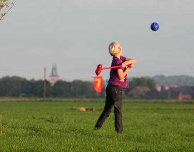 Ein Kind hat einen Ball mit einem Schläger weggestoßen. Es steht auf einer grünen Wiese. Der Ball ist Blau und fliegt durch die Luft.