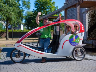 Eine Fahrrad-Rikscha, der Fahrer Winkt in die Kamera und in der Rikscha sitzen zwei Frauen