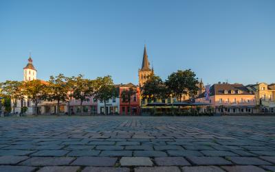 Großer Platz mit Kopfsteinpflaster und Bäumen, im Hintergrund eine Statue des Hl. Viktor sowie historische Gebäude und Kirchtürme bei klarem Himmel