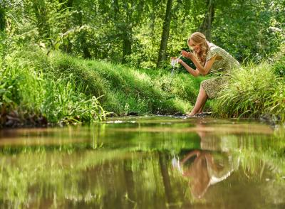 Eine Frau sitzt an einem kleinen Fluss inmitten einer grünen Landschaft . Sie hat ihre Füße im Wasser und lässt das Wasser aus dem Fluss durch ihre Hände gleiten.