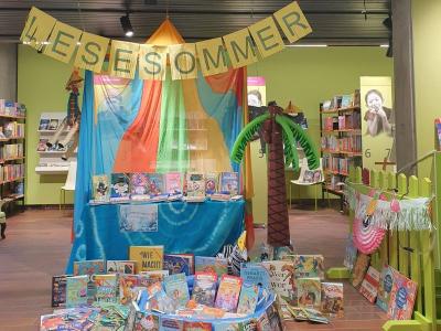 Bunte Sommer-Leseecke in einer Bibliothek mit dem Schriftzug 'Lesesommer'. Der Bereich ist dekoriert mit bunten Tüchern, einem aufblasbaren Palmenbaum und zahlreichen Kinderbüchern, die auf einem Tisch und am Boden ausgestellt sind.